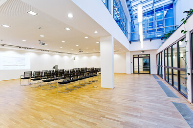 Foto des geräumigen und offenen Konferenzraums im Atrium mit Kinobestuhlung für ca. 90 Personen, Rednerpult und Beamer. Durch große Fenster im überdachtem Bereich zeigt sich das Atrium lichtdurchflutet. Pflanzen auf einer Galerie sorgen für eine angenehme Atmosphäre.